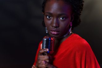 Mulher vestida de vermelho canta ao microfone diante de um fundo escuro.